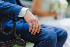 Man, groom in a blue suit sitting on a bench. Close-up. A hand with a ring. Bride with flowers in the background.